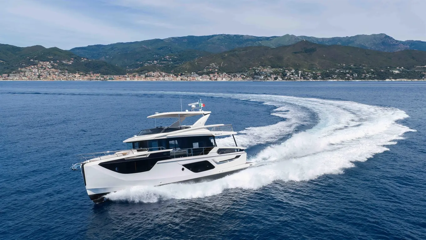 A luxury yacht speeds across a calm sea, leaving a wake, with a coastal town and mountains in the background.